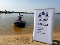 Jezioro, na nim policyjna motorówka ze stojacym w niej policjantem. Na pierwszym planie na plazy baner z napisem komenda miejska policji w Gliwicach.