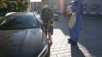 dziecko w towarzystwie policjanta wręcza rysunek kierujacej samochodem (zatrzymanej na ulicy). Po prawej stoi człowiek w stroju policyjnej maskotki psa sznupka.