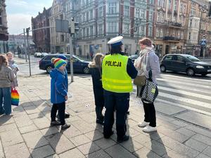 Na zdjęciu rejon przejścia dla pieszych. Policjantka rozmawia z pieszą. wokół stoją dzieci szkolne i przysłuchują się....